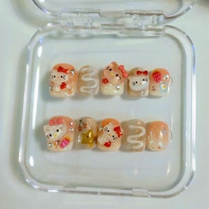 Könnte beinhalten: Ein Set aus zehn künstlichen Nägeln in einer transparenten Kunststoffbox. Die Nägel weisen verschiedene Designs auf, darunter Hello Kitty-Charaktere, Erdbeeren und Glitzerakzente. Die Farbpalette umfasst Rosa, Weiß und Gold.