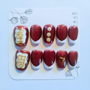 May include: A set of press-on nails in a variety of sizes. The nails are primarily burgundy with white tips. Some nails feature gold lettering that spells "ARMY" and "BTS", while others have gold accents or the number "7".