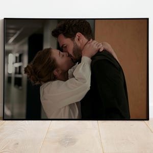 May include: A rectangular framed print featuring a photographic scene of two individuals kissing. One individual wears a long-sleeved white button-down shirt, while the other wears a dark black shirt. The scene is set in an indoor hallway environment.