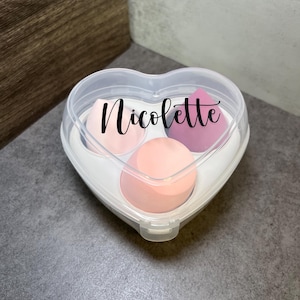 May include: A clear heart-shaped container with three pink makeup sponges. The container has the name "Nicolette" written on it.