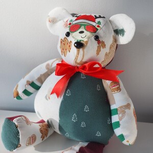 May include: A handmade teddy bear with a festive design. The bear features a white head with a Santa hat and sunglasses, a green waffle-textured body with white Christmas trees, and red accents. A red ribbon is tied around the neck.