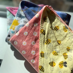 May include: Three folded fabric items with different patterns. The front item is yellow with black and yellow bees. The middle item is pink with white daisies. The back item is blue and white with a cartoon animal.