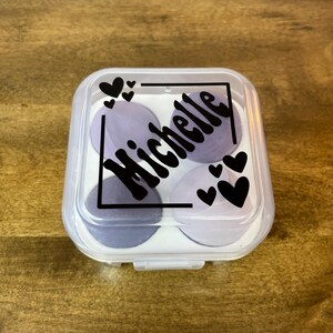 May include: A clear, square container with a hinged lid, holding four circular makeup sponges in shades of purple and gray. The lid features the name "Michelle" in a black, stylized font, along with black heart accents.