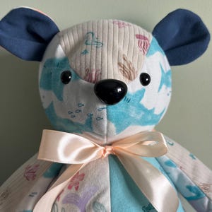 May include: A handmade plush bear with a patchwork design. The bear features a black nose, black button eyes, and navy blue ears. The body is made of various fabrics with sea-themed patterns, including sharks and coral. A light pink ribbon is tied around the neck.