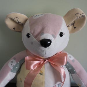 May include: A handmade plush toy in the shape of a mouse, featuring a pink and white face, large black eyes, and a black nose. The ears are beige with a whimsical design. A pink satin bow adorns the neck. The body is a patchwork of pastel fabrics.