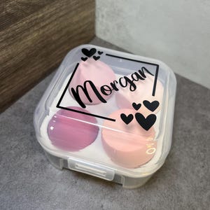 May include: A clear plastic container with a hinged lid. The container has three pink makeup sponges inside. The lid has a black and white design with the name "Morgan" and hearts.