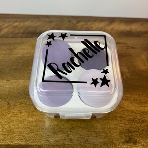May include: A clear plastic container with a lid, containing four purple makeup sponges. The lid has a black label with the name "Rochelle" and stars printed on it.