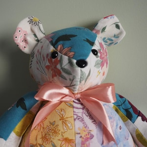 May include: A handmade teddy bear with a patchwork design. The bear's head is teal and white with floral patterns, and the ears are white with pink flowers. It has a pink satin bow and black button eyes.