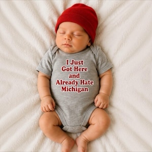 May include: A grey short-sleeved baby bodysuit featuring the red text I Just Got Here and Already Hate Michigan, paired with a solid red knit beanie. The garment is made of a soft fabric and is designed for infants, showcasing a clear graphic print.