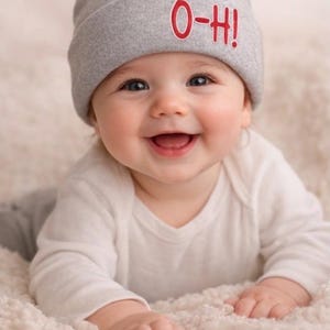 May include: A happy baby wearing a grey beanie hat with the red text "O-Hi!". The baby is dressed in a white onesie and lying on a soft, white surface.