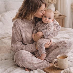 Op de afbeelding: Een moeder en baby in bijpassende tie-dye pyjama's. De moeder kust het voorhoofd van de baby. Een mok koffie staat op een houten dienblad op het bed. De scène is zacht en gezellig.