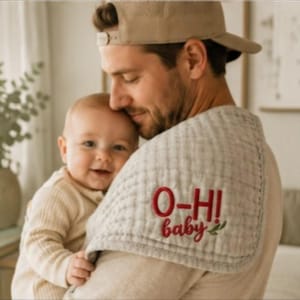 Könnte beinhalten: Ein hellgraues, strukturiertes Spucktuch mit dem gestickten roten Text O-HI und dem Wort baby in roter Schreibschrift, akzentuiert durch eine kleine grüne Blattgrafik. Das Tuch hat eine abgerundete Ecke und einen genähten Rand.