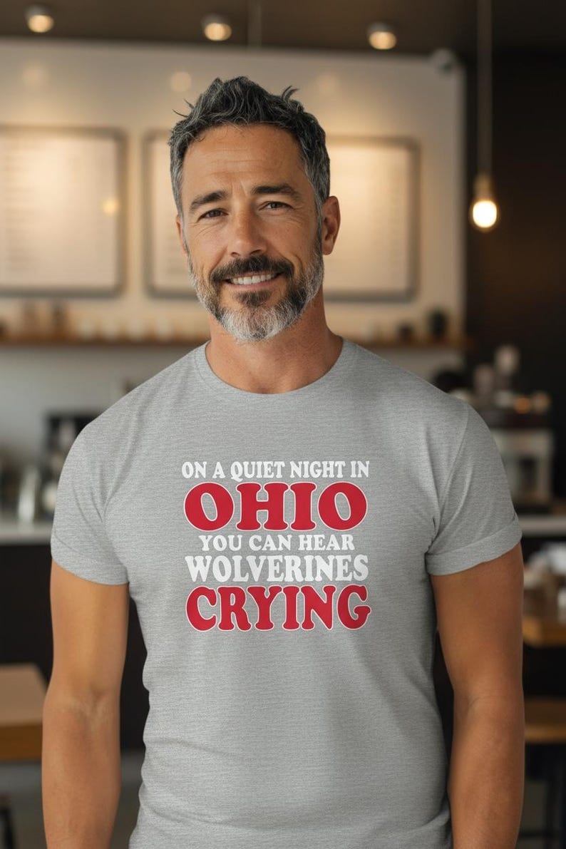 May include: A light grey t-shirt with the text "ON A QUIET NIGHT IN OHIO YOU CAN HEAR WOLVERINES CRYING" in red and white. The shirt is worn by a man with grey hair and a beard.