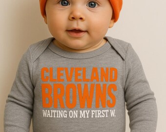 May include: A baby wearing an orange knit cap and a gray onesie. The onesie has the words "CLEVELAND BROWNS" in orange block letters, with the phrase "WAITING ON MY FIRST W" below. The baby has blue eyes.