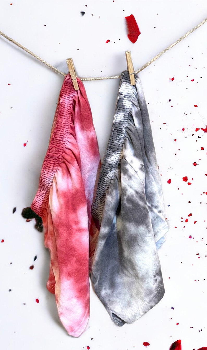 May include: Two tie-dye fabric panels hanging on a clothesline with wooden clothespins. The panel on the left is pink and white, and the panel on the right is gray and white.
