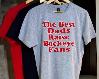 May include: Three t-shirts hanging on wooden hangers. The first is red, the second is black, and the third is gray. The gray t-shirt has the text "The Best Dads Raise Buckeye Fans" in red letters.