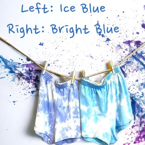 May include: Two pairs of tie-dye shorts hanging on a clothesline. The shorts on the left are a lighter ice blue color, while the shorts on the right are a brighter blue. The text "Left: Ice Blue" and "Right: Bright Blue" is written above the shorts.