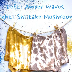 May include: Two pairs of tie-dye shorts hanging on a clothesline. The left pair is yellow and white with the text "Amber Waves". The right pair is brown and white with the text "Shiitake Mushroom".