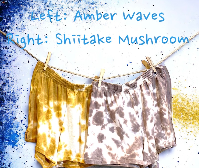 May include: Two pairs of tie-dye shorts hanging on a clothesline. The left pair is yellow and white with the text "Amber Waves". The right pair is brown and white with the text "Shiitake Mushroom".