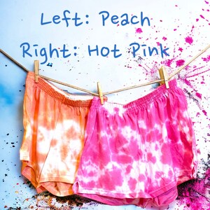 May include: Two pairs of tie-dye shorts hanging on a clothesline. The shorts on the left are peach colored with white tie-dye. The shorts on the right are hot pink with white tie-dye. Left: Peach. Right: Hot Pink.