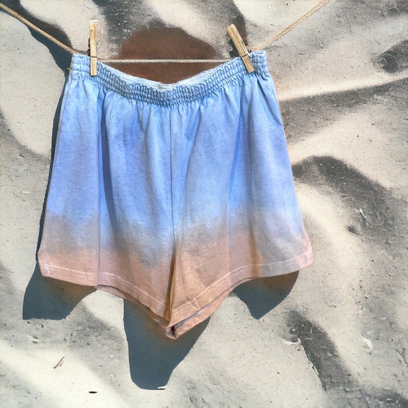 May include: A pair of blue and peach tie-dye shorts hanging on a clothesline against a sandy background.