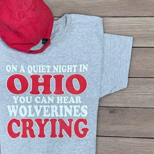 May include: A grey t-shirt with the text "ON A QUIET NIGHT IN OHIO YOU CAN HEAR WOLVERINES CRYING" in red and white. A red baseball cap is placed on the shirt. The shirt is made of a soft material.