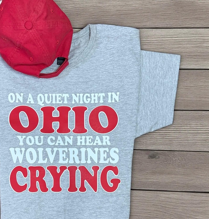 May include: A grey t-shirt with the text "ON A QUIET NIGHT IN OHIO YOU CAN HEAR WOLVERINES CRYING" in red and white. A red baseball cap is placed on the shirt. The shirt is made of a soft material.