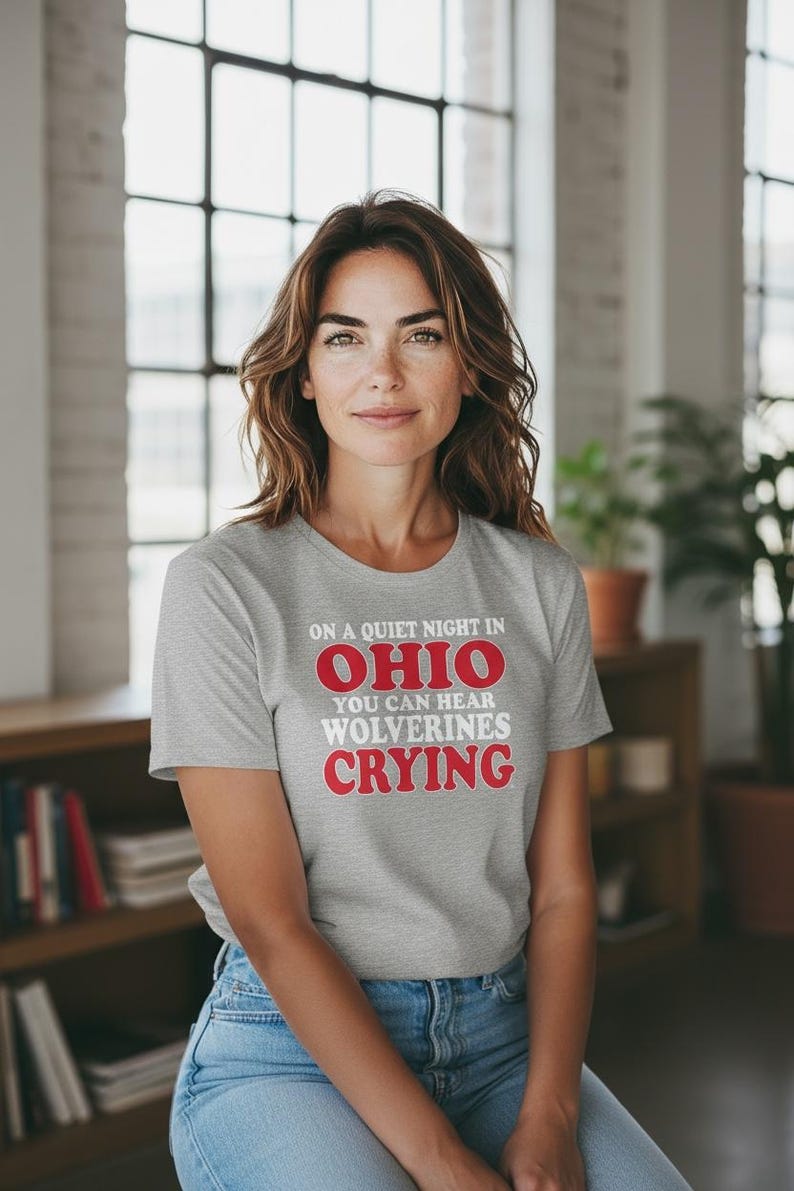 May include: A woman in a grey t-shirt with the words "ON A QUIET NIGHT IN OHIO YOU CAN HEAR WOLVERINES CRYING" in red and white. She is wearing blue jeans. The background includes a window and a bookshelf.