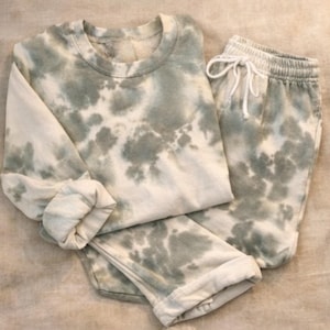 May include: A tie-dye lounge set in green and white. The set comprises a crew neck sweatshirt and drawstring sweatpants. The fabric has a soft, cloud-like pattern. The cuffs and waistband are ribbed.