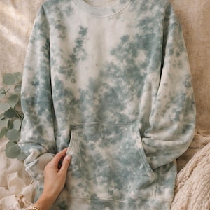 Może przedstawiać: Jasnozielona i biała bluza z efektem tie-dye z kieszenią typu kangur. Bluza z długim rękawem ma okrągły dekolt i luźny krój. Tkanina ma miękki, teksturowany wygląd. Bluza jest prezentowana na neutralnej powierzchni.