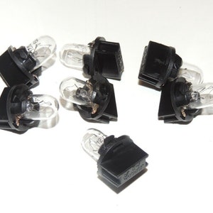 Puede incluir: Un conjunto de ocho bombillas negras y transparentes. Las bombillas tienen una bombilla de vidrio transparente y una base de plástico negro. Las bombillas están dispuestas en círculo sobre una superficie blanca.