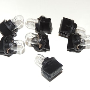 Puede incluir: Un conjunto de ocho bombillas negras y transparentes. Las bombillas tienen una bombilla de vidrio transparente y una base de plástico negro. Las bombillas están dispuestas en círculo sobre una superficie blanca.