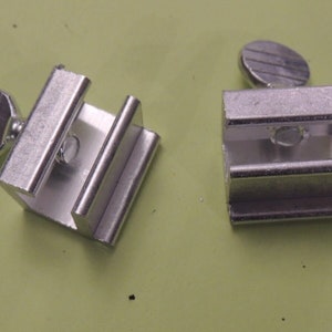 May include: Two small, silver metal clamps with screw-on knobs. The clamps are rectangular with a channel for holding objects. The knobs are round and have a ridged texture.