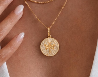 May include: A layered gold necklace set featuring a delicate chain, a twisted rope chain, and a longer chain with a round pendant. The pendant displays a dragonfly motif set against a background of small clear stones. Two thin gold rings are worn.
