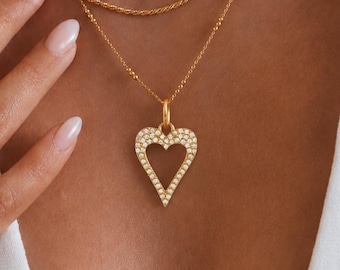 May include: A layered gold necklace set featuring a delicate chain, a rope chain, and a longer chain with a heart-shaped pendant encrusted with small clear stones. Two thin gold rings are worn on the fingers. The text ONE SOURCE is visible.