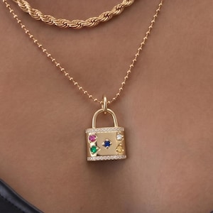 May include: A layered gold-tone necklace set consisting of three chains: a thick rectangular link chain, a twisted rope chain, and a delicate ball chain. The longest chain features a rectangular padlock pendant adorned with small colorful gemstones.