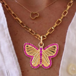 May include: A gold-toned necklace featuring a large butterfly pendant with bright pink stones lining the wings and body, suspended from a thick chain, layered with a delicate gold chain featuring a small heart-shaped pendant.