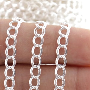 Puede incluir: Tres hebras de cadena de plata, cada una con un diseño delicado de eslabones redondeados. La cadena se muestra sobre una mano, mostrando su tamaño y textura.