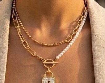 Puede incluir: Un collar dorado de varias capas que combina una cadena fina, una cadena de eslabones gruesos y una hilera de perlas blancas. El collar incluye un cierre de barra en T dorado y un colgante de candado rectangular incrustado con piedras claras.