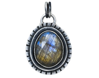 May include: A silver pendant featuring a blue and gold labradorite gemstone set in a silver bezel. The pendant has a textured, circular border.