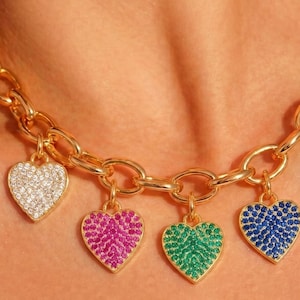 May include: A gold-toned chain necklace featuring four heart-shaped charms. Each heart is encrusted with small, sparkling stones in white, pink, green, and blue. The necklace has a thick, interlocking link design and rests around the neck area.