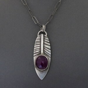 May include: A silver necklace with a pendant shaped like a feather. The feather has a purple gemstone in the center.