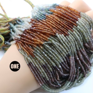 Peut inclure: Un brin de perles de pierres précieuses facettées dans des tons de brun, vert et gris. Les perles sont petites et de taille uniforme, et elles sont enfilées sur un cordon fin. Le brin mesure environ 45 cm de long. Le mot "ONE" est écrit en noir sur un cercle blanc dans le coin inférieur gauche de l'image.