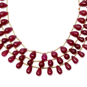 May include: A strand of ruby red gemstone beads in a teardrop shape, strung on gold wire. The beads are arranged in three rows, with tassels on each end.
