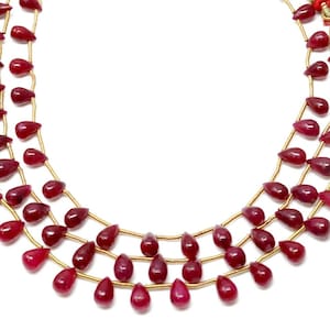 May include: A strand of ruby red gemstone beads in a teardrop shape. The beads are strung on gold wire and have a tassel on each end.