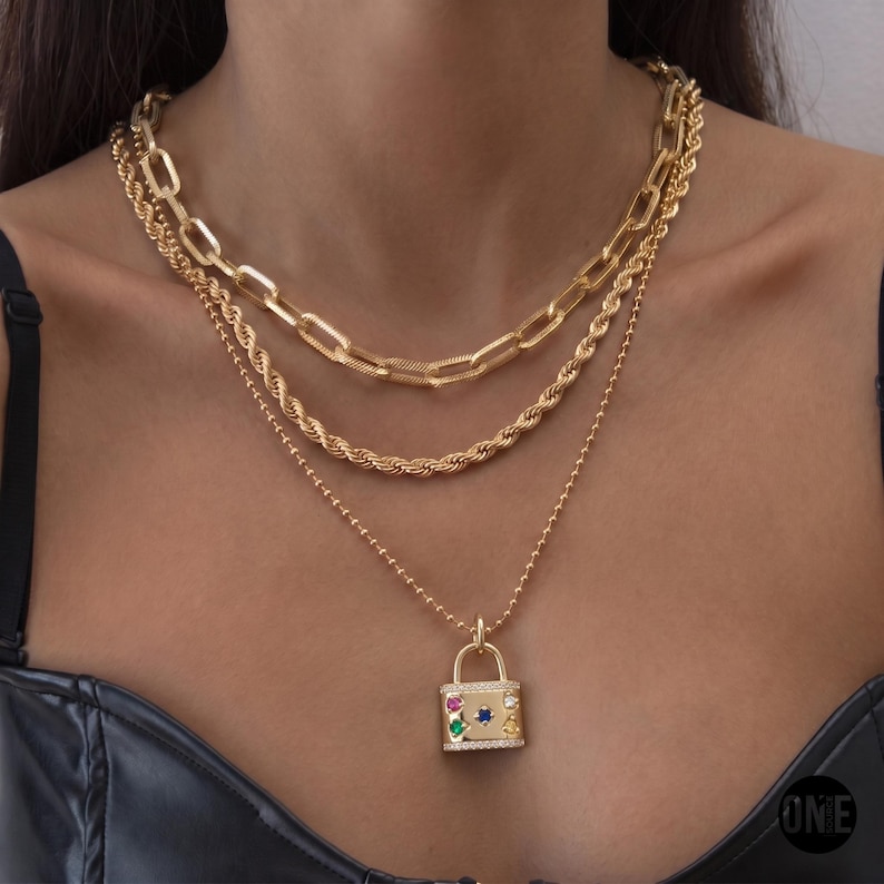 May include: A layered gold-tone necklace set consisting of three chains: a thick rectangular link chain, a twisted rope chain, and a delicate ball chain. The longest chain features a rectangular padlock pendant adorned with small colorful gemstones.