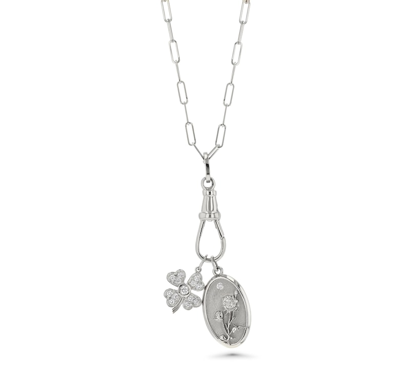 May include: A silver-toned necklace with a paperclip chain design. The necklace features a clasp with two charms: a four-leaf clover and an oval pendant with a floral design, both adorned with small, sparkling stones.