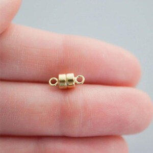 May include: A small, gold-colored magnetic clasp for jewelry making. The clasp is cylindrical with two small loops on either end. The clasp is shown against a blurred background.