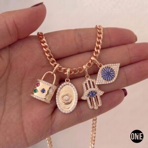 May include: A rose gold chain necklace featuring four charms: a padlock with multi-colored stones, an oval medallion with a central stone, a hamsa hand with a blue stone, and an eye-shaped charm with blue stones, all accented with clear crystals.