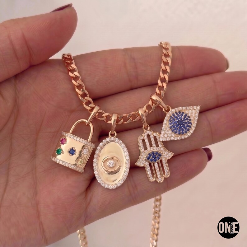 May include: A rose gold chain necklace featuring four charms: a padlock with multi-colored stones, an oval medallion with a central stone, a hamsa hand with a blue stone, and an eye-shaped charm with blue stones, all accented with clear crystals.